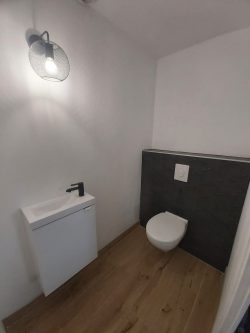 WC suspendu avec bâti-support encastré et lave-mains compact dans un espace sanitaire optimisé. Le sol effet bois et la finition murale noire texturée apportent une touche élégante et moderne. Réalisation WT TOUS TRAVAUX à Montbéliard.